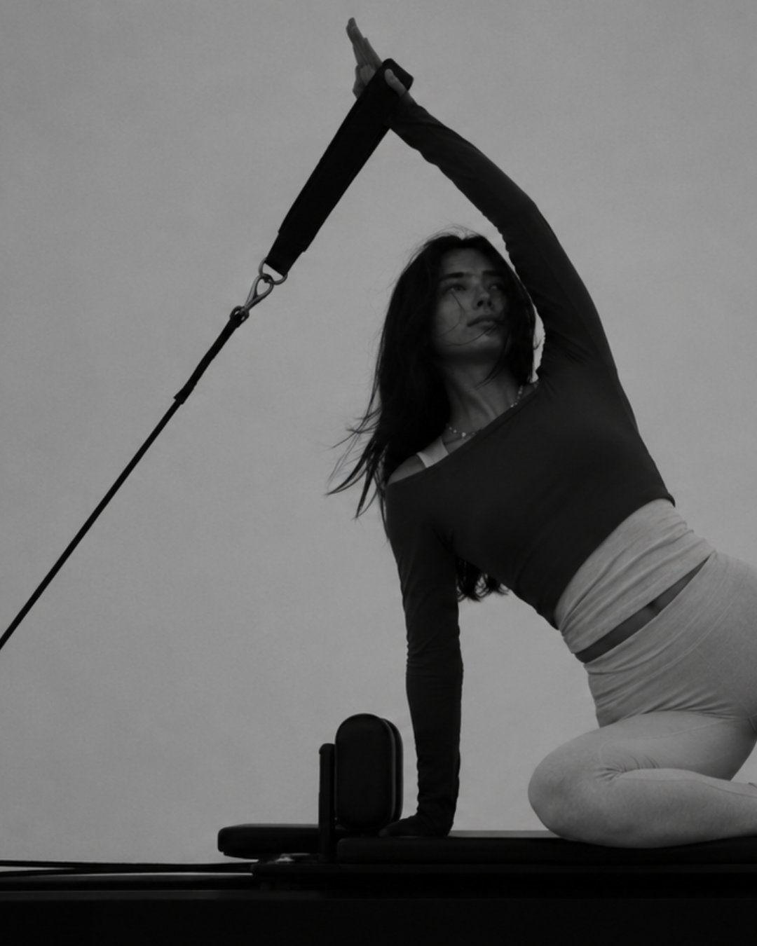 Black-and-white image of a flowhouse client kneeling on the FlowFormer in a slow side stretch, holding a strap overhead.