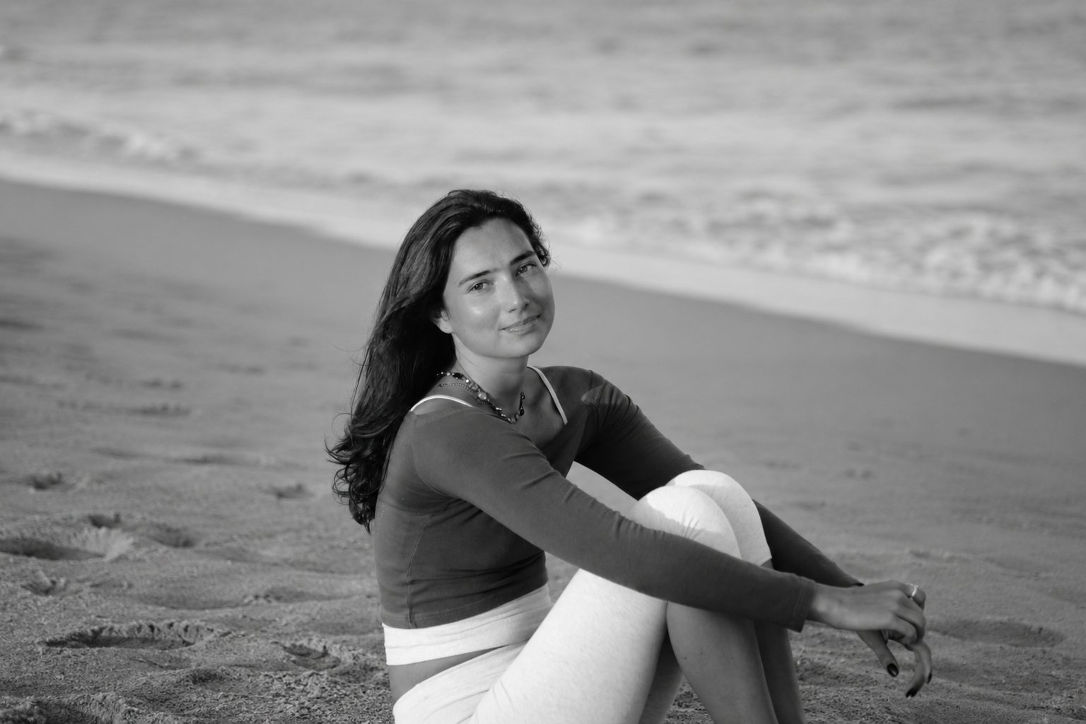 Black-and-white image of a flowhouse client seated quietly on the beach, hugging her knees.
