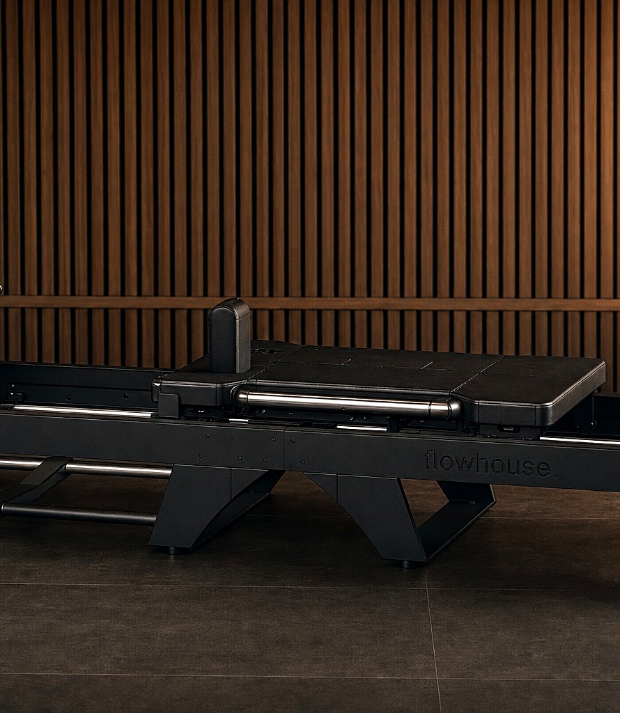 The custom flowhouse FlowFormer reformer — matte-black frame with the flowhouse wordmark on the side rail, against a warm walnut wood-slat wall.